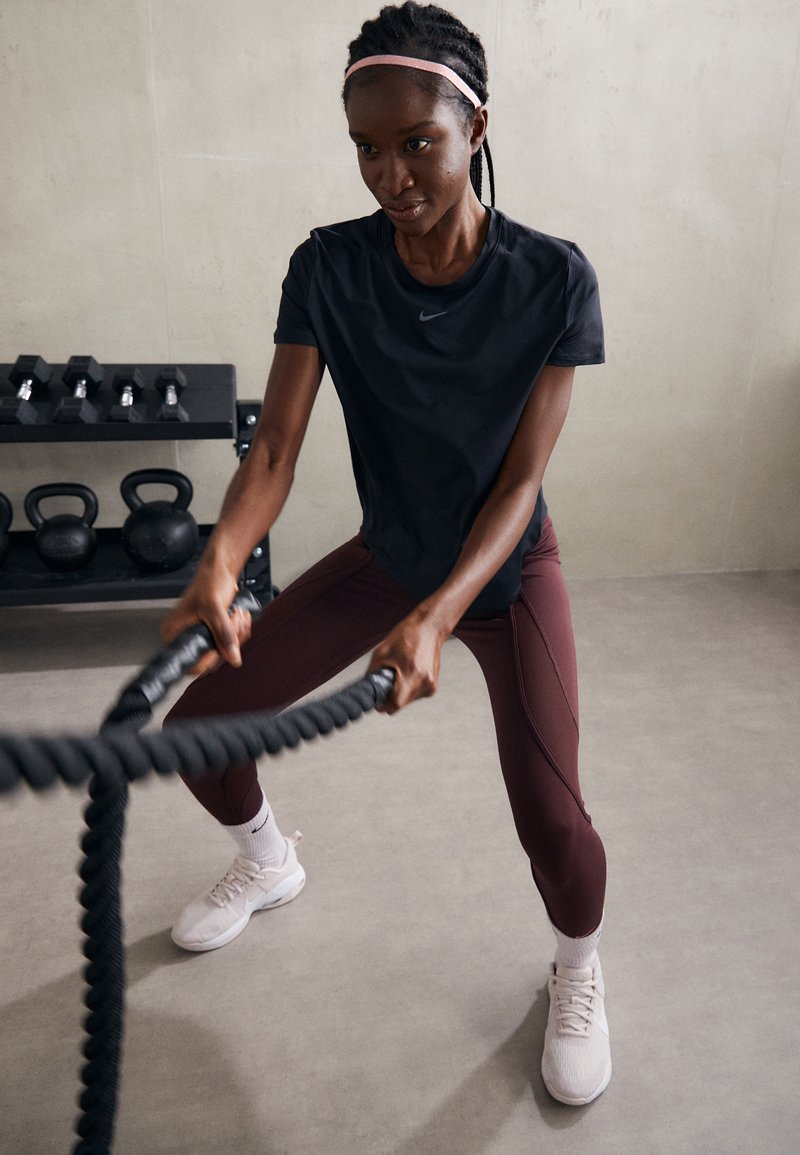 Schwarzes sportliches T-Shirt, kastanienbraune Leggings mit Nahtakzenten und weiße Sneaker. Die Person trainiert mit dicken schwarzen Battle Ropes.