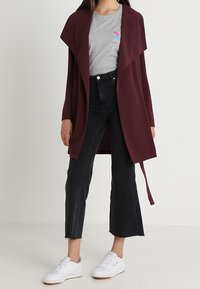 Burgundowa narzutka z otwartym przodem, długimi rękawami i wiązaniem. W połączeniu z szarym t-shirtem, czarnymi, wysokimi dżinsami ściętymi w kostkach i białymi sneakersami.