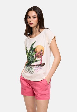 Joven con cabello largo y oscuro, lleva una camiseta gráfica beige con un barco y plantas, combinada con pantalones cortos de color rosa brillante, manos en los bolsillos.