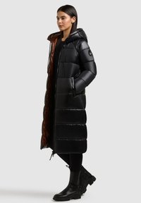 Hosszú fekete pufi kabát fényes külsővel, vízszintes varrással és barna béléssel. Kapucnival és cipzáras zsebekkel rendelkezik. Fekete csizmák.