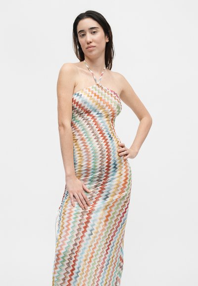Γυναίκα που στέκεται με το ένα χέρι στη μέση, φοράει αμάνικο φόρεμα λαιμόκοψης με πολύχρωμο ζιγκ-ζαγκ μοτίβο σε λευκό φόντο.