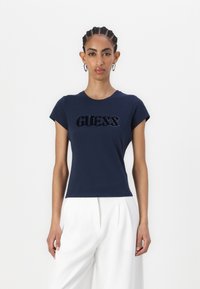 Tamsiai mėlynas prigludęs marškinėliai, pagaminti iš minkštos medvilnės, su blizgiu „GUESS“ logotipu tekstūruotame dizaino. Trumpomis rankovėmis ir apvali iškirpte.