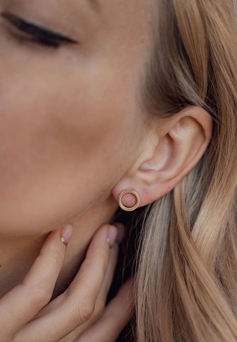 Boucle d'oreille en or en forme de cercle portée à l'oreille, finition métallique texturée, design simple ; un arrière-plan flou met en valeur la forme de la boucle.