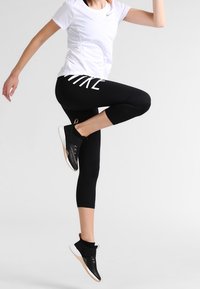 Maglietta sportiva bianca Nike, leggings alla caviglia neri con logo bianco e scarpe da ginnastica nere con dettagli testurizzati, in una posa dinamica di salto.
