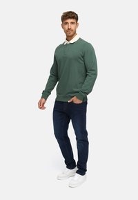 Groene pullover sweatshirt met een witte kraag, gecombineerd met donkerblauwe jeans en witte sneakers. Heeft ribgebreide boorden en een ronde hals.