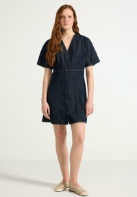 Jeansklänning med V-ringning, korta ärmar och knappar fram. Mörkblå färg med synlig sömnad och en figursydd midja.