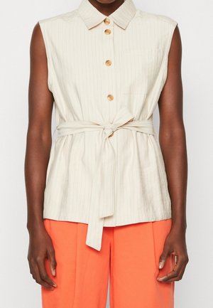 Chemise crème sans manches à rayures verticales, boutonnée devant, ceinture nouée à la taille, portée avec un pantalon orange vif sur une personne debout.
