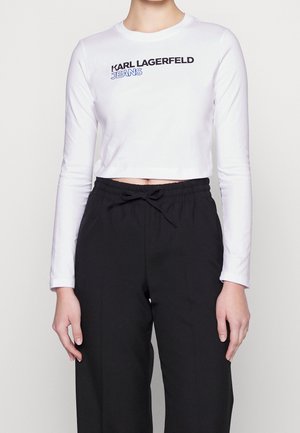Femme portant un crop top blanc à manches longues avec l'inscription « KARL LAGERFELD JEANS » et un pantalon noir à cordon, debout devant un fond uni.