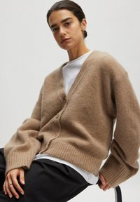 Bruine wollen cardigan met een V-hals, knopen aan de voorkant en geribbelde manchetten. Draag het over een wit T-shirt met een zachte, textuur.