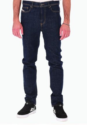 Homme portant un jean en denim bleu foncé et des baskets noires avec des semelles blanches, debout devant un fond blanc.