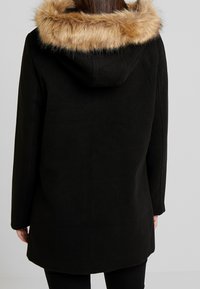 Sort frakke med en fluffy, lys brun pelsforet hætte. Designet er strømlinet, med en lige skæring og lange ærmer.