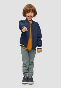 Marine bomberjack met witte accenten, oranje graphic t-shirt, lichtgrijze broek en marineblauwe schoenen met rode details; kind dat glimlacht en wijst.