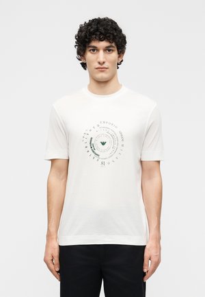 Άντρας με σγουρά μαλλιά που φοράει ένα λευκό T-shirt με κυκλικό μαύρο και πράσινο λογότυπο Emporio Armani, σε συνδυασμό με μαύρο παντελόνι.