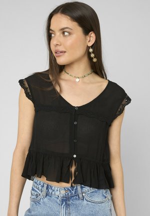 Jeune femme aux longs cheveux foncés porte un chemisier noir transparent boutonné avec des volants, un jean bleu, des boucles d’oreilles en forme de soleil et un collier de perles.