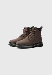 Botas de cuero marrón con cordones, con un cuello acolchado y suelas de goma negra, inclinadas ligeramente hacia la derecha sobre un fondo claro y liso.