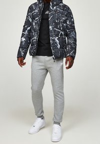 Muž v černo-bílé bundě s abstraktním vzorem, černém tričku, šedých teplácích a bílých teniskách stojící na neutrálním pozadí.