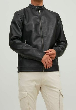 Persona che indossa una giacca di pelle nera con cerniera e pantaloni cargo beige chiaro con le mani intrecciate davanti.