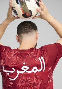 Jersey punah merah dengan pola geometris rumit dan tulisan Arab putih "المغرب". Individu tersebut memegang bola sepak berpola di atas kepala mereka.