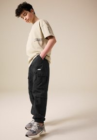 Béžové tričko s čiernym textom, skombinované s čiernymi cargo nohavicami s viacerými vreckami. Šedé tenisky s bielymi šnúrkami dotvárajú outfit.