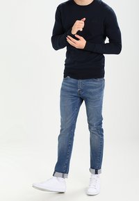 Námořnicky modrý pletený svetr s kulatým výstřihem, světle modré džíny s rovnými nohavicemi a bílé tenisky. Otočené manžety na džínách, na svetru minimální detail s logem.