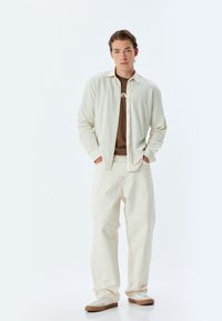 Camisa de botón color crema sobre una camiseta marrón, combinada con pantalones anchos blancos y zapatillas beige, con detalles de costura contrastantes.