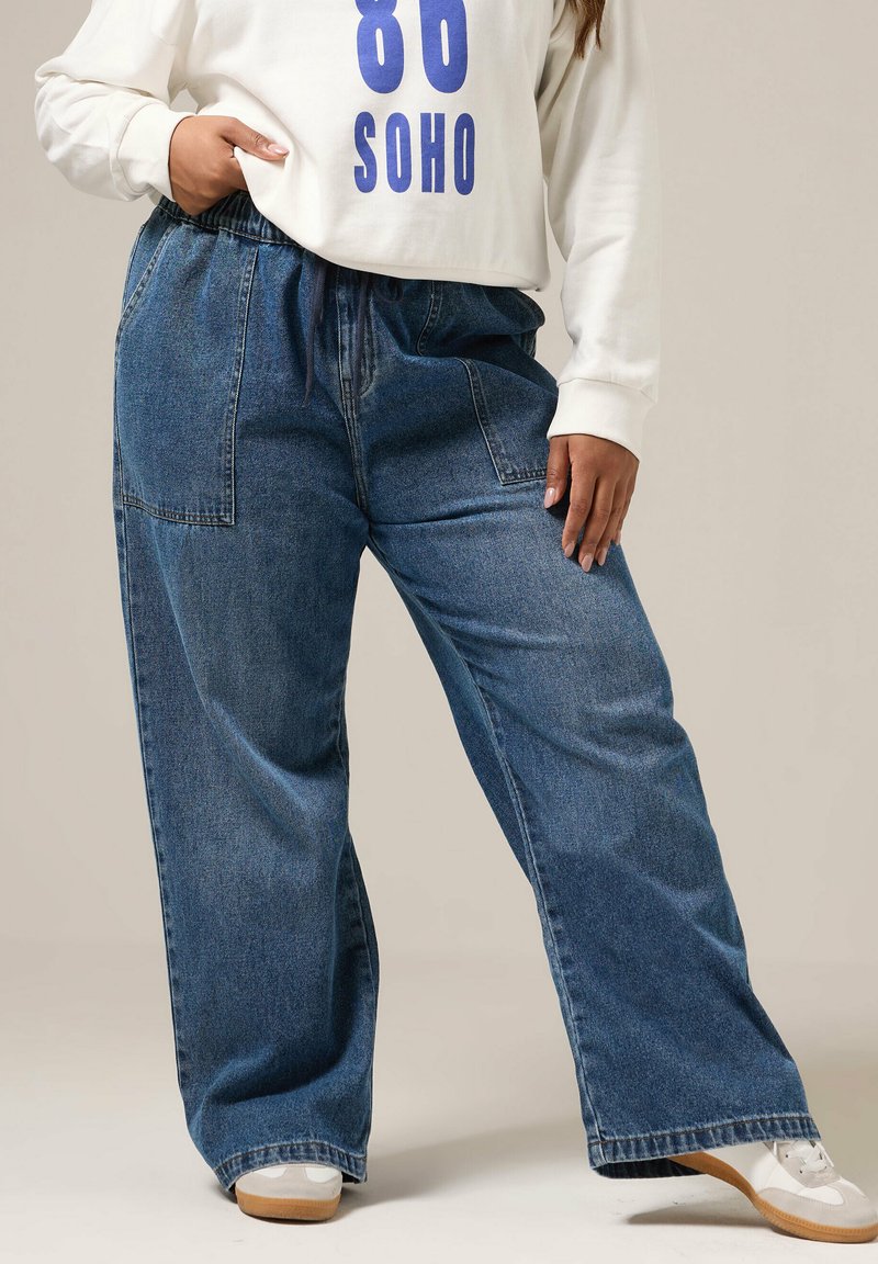 Persona che indossa un paio di jeans in denim blu a vestibilità ampia con grandi tasche anteriori, una felpa bianca con il testo blu "86 SOHO" e sneakers bianche.