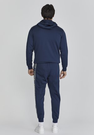 Sudadera y pantalones de chándal en un suave tejido navy, con puños acanalados y una franja con logo en los pantalones. El diseño es atlético y ajustado.