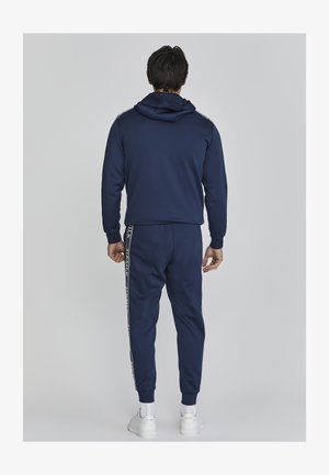 Jūras spēku kapuce un bikses no mīkstas auduma, ar ribotām aprocēm un logo svītru gar biksēm. Dizains ir sportisks un pieguļošs.