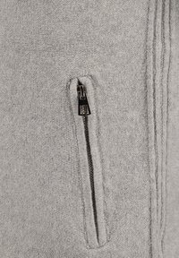 Tascone grigia con zip su un capo di abbigliamento testurizzato, decorata con un tirante della zip in argento dotato di una linguetta rettangolare e con dettagli di cucitura sottili sul lato.
