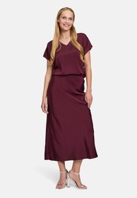 Burgunderfarbendes Satin-Kleid mit V-Ausschnitt, kurzen Ärmeln und einer Länge bis zur Mitte der Wade. Es hat eine lockere Passform und eine glatte Textur, kombiniert mit silbernen Absätzen.