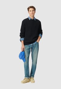 Czarny sweter o teksturze nakładany na jasnoniebieską koszulę dżinsową, w komplecie z jasnoniebieskimi dżinsami i beżowymi butami. Trzyma niebieską czapkę.