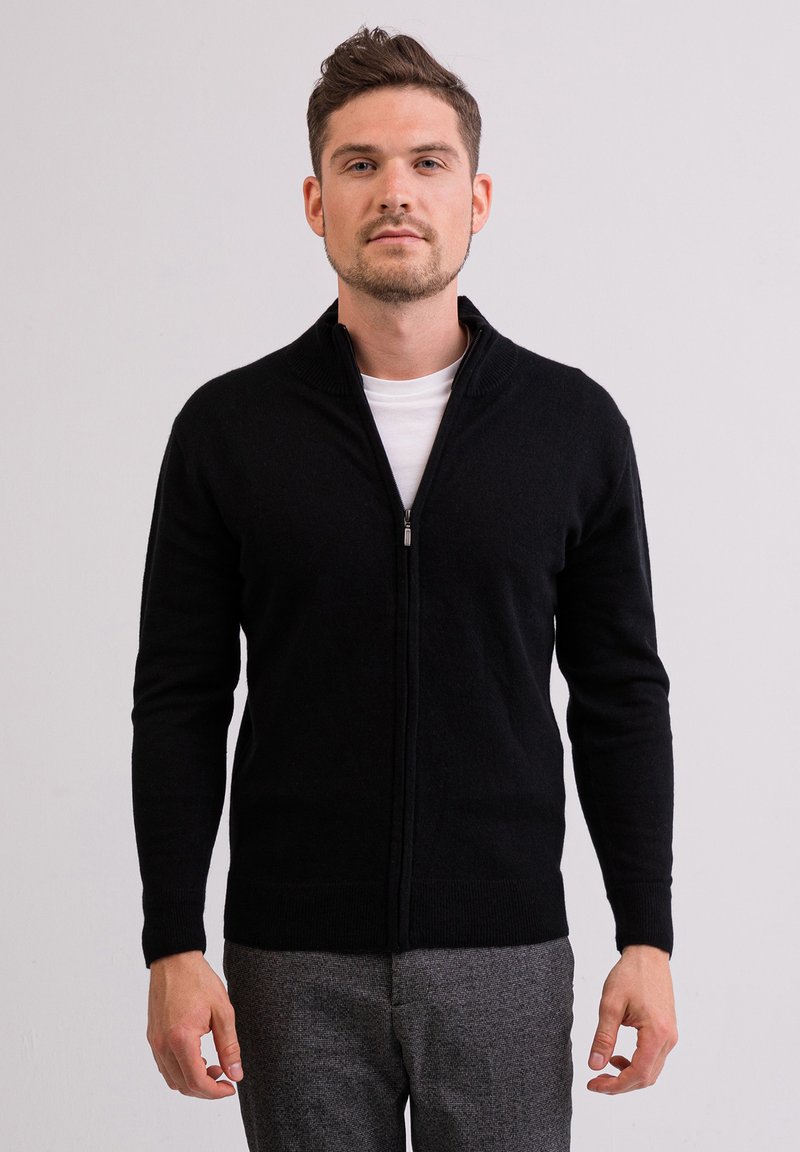 Mustapukuinen vetoketjullinen neule, jossa on korkea kaulus, neulottu pinta ja pitkät hihat, päällä valkoinen paita. Istuu tiiviisti vartalon ympärille.