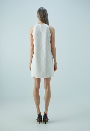 Robe sans manches blanche avec un motif texturé, comportant un trou de serrure à l'arrière. Portée avec des chaussures à talons noires, mettant en valeur un design minimaliste.