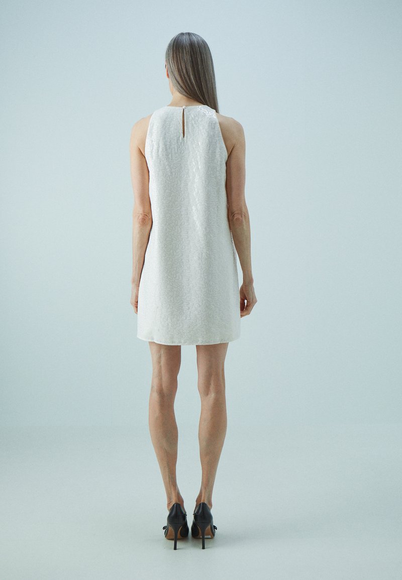 Robe sans manches blanche avec un motif texturé, comportant un trou de serrure à l'arrière. Portée avec des chaussures à talons noires, mettant en valeur un design minimaliste.