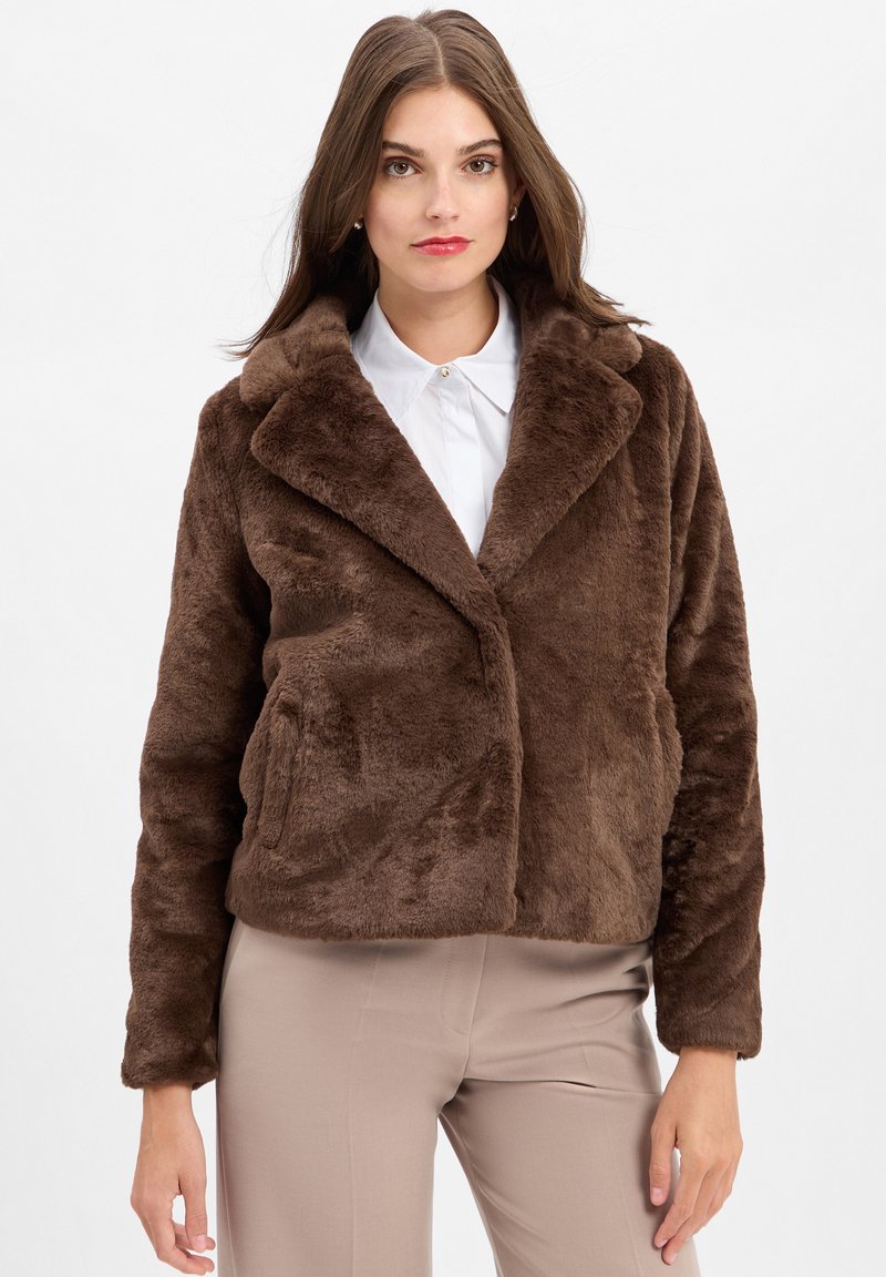 Bruine faux-bont jas met een sjalkraag, cropped ontwerp en zijzakken, gedragen over een wit overhemd met een kraag en beige broek.