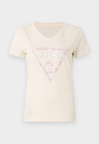 T-shirt en coton beige à manches courtes avec un col en V et un logo en triangle pailleté rose portant le texte "GUESS" et des accents embellis.