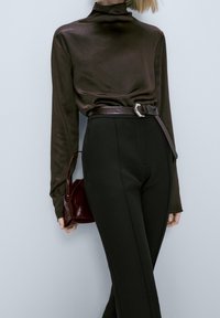 Haut à col roulé en satin brun associé à un pantalon noir taille haute, maintenu par une ceinture en cuir foncé. Un petit sac à main bordeaux complète le look.