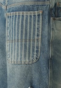 Niebieskie szorty dżinsowe z wyjątkową tylną kieszenią ozdobioną pionowymi paskami i kontrastującym przeszyciem, podkreślające teksturowaną tkaninę i swobodny styl.