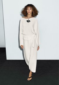 Long, loose-fitting cream dress made of soft material, featuring a wrap tie at the waist. Worn with a black heart-shaped necklace and sandals.