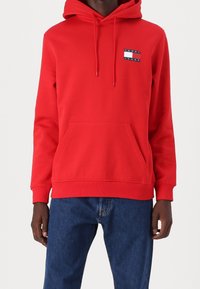 Felpa rossa in cotone con tasca a marsupio, cappuccio con laccetto e un patch con logo che presenta un design a strisce blu, bianche e rosse sul petto.