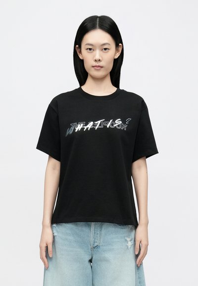 Μαύρο βαμβακερό t-shirt με κοντά μανίκια που φέρει αντανακλαστικό κείμενο "WHAT IS?" στο μπροστινό μέρος. Συνδυάζεται με ανοιχτό μπλε τζιν παντελόνι με σκισίματα.