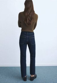 Femme aux longs cheveux bruns portant un pull marron, un jean bleu foncé et des chaussures noires, debout les bras croisés face à un mur blanc.