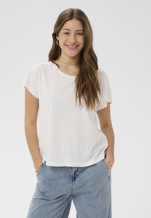 Mladá žena s dlhými hnedými vlasmi, oblečená v bielej tričku s krátkymi rukávmi a modrých džínsoch, usmievajúca sa s rukami vreckách na jednoduchom pozadí.