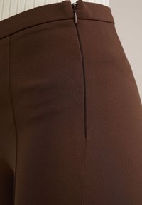 Pantalones de tiro alto marrones con una textura suave, detalle de cremallera lateral y un ajuste a medida, ideales para ocasiones casuales o formales.