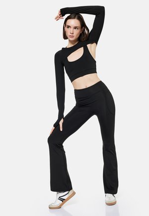 Donna con crop top nero con ritagli, pantaloni neri a vita alta e gamba svasata, e sneaker bianche posa con un braccio alzato e una gamba piegata.