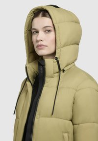 Jachetă pufoasă de culoare măsline deschisă, cu glugă, având un fermoar în față, accente negre, textură matlasată și șnururi pentru ajustarea potrivirii.