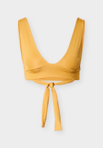 Κίτρινο κορμάκι από απαλό ύφασμα με βαθύ V-λαιμόκοψη, φαρδιές τιράντες και δεμένη λεπτομέρεια στη μέση. Απλό σχέδιο, χωρίς μοτίβα ή μεταλλικά στοιχεία.