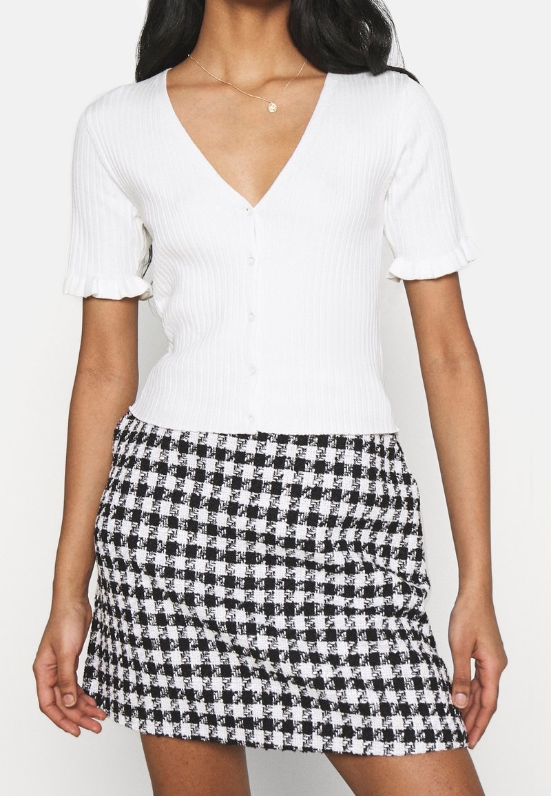 Femme portant un haut blanc à manches courtes côtelé avec boutons et poignets volantés, ainsi qu'une mini-jupe pied-de-poule noir et blanc.
