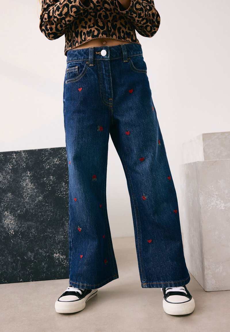 Personne portant un jean large bleu foncé avec une petite broderie rouge cœur et rose, des baskets noires et blanches, et un top court imprimé léopard.