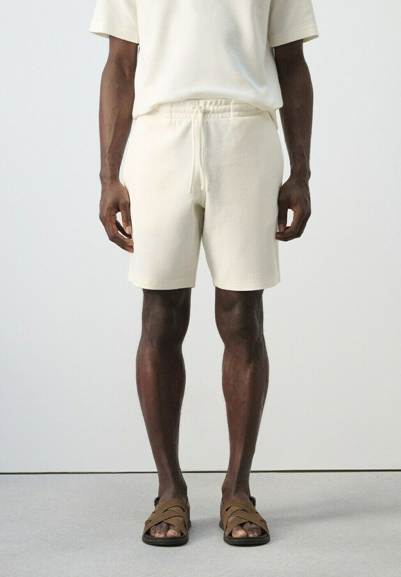 Homme portant un short texturé blanc cassé avec taille à cordon de serrage, un haut à manches courtes assorti, et des sandales marron croisées, debout sur un sol gris.